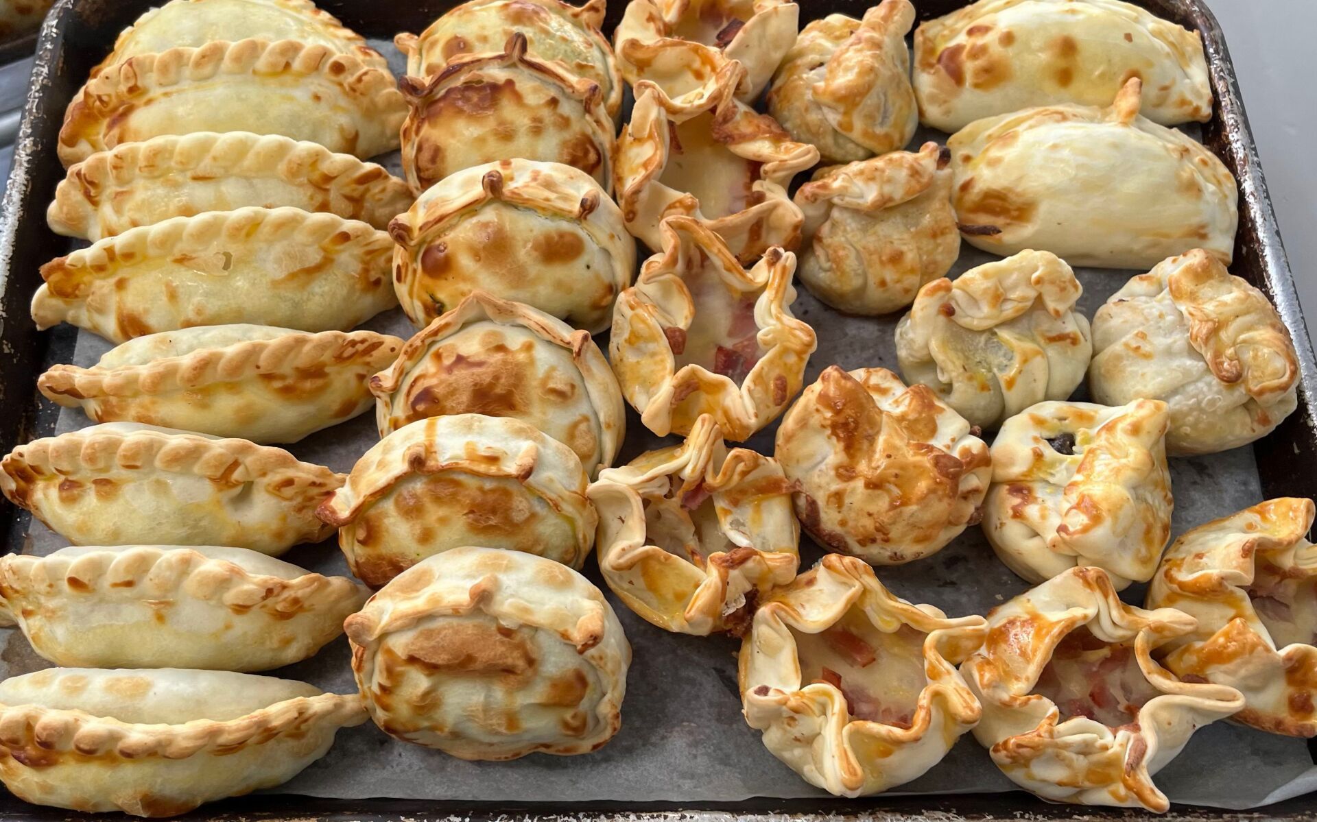 Tray of golden-brown, baked empanadas with crimped edges and various shapes.