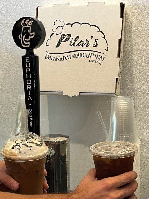 Two iced coffees held near a beer tap and a Pilar's Empanadas box.