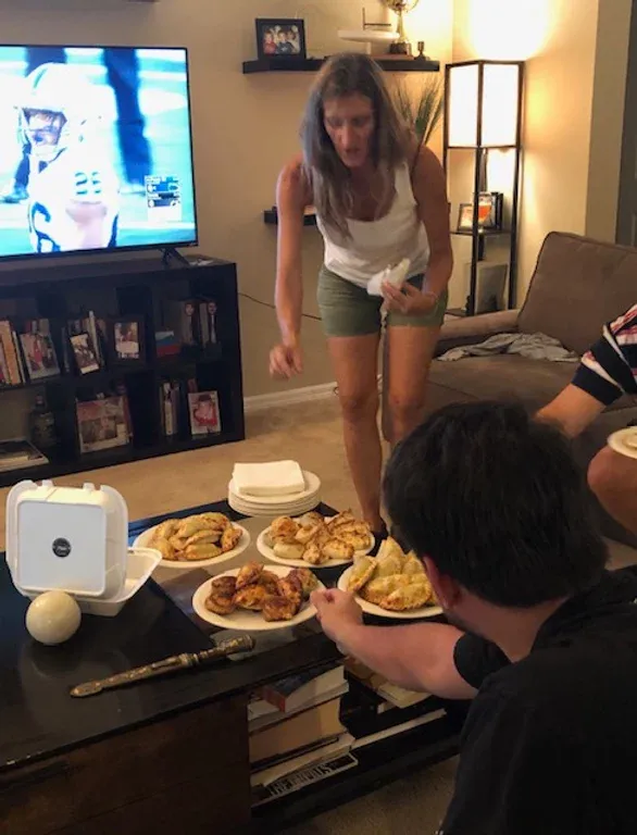 Woman gestures at food on coffee table, man reaches for it. TV showing football game.