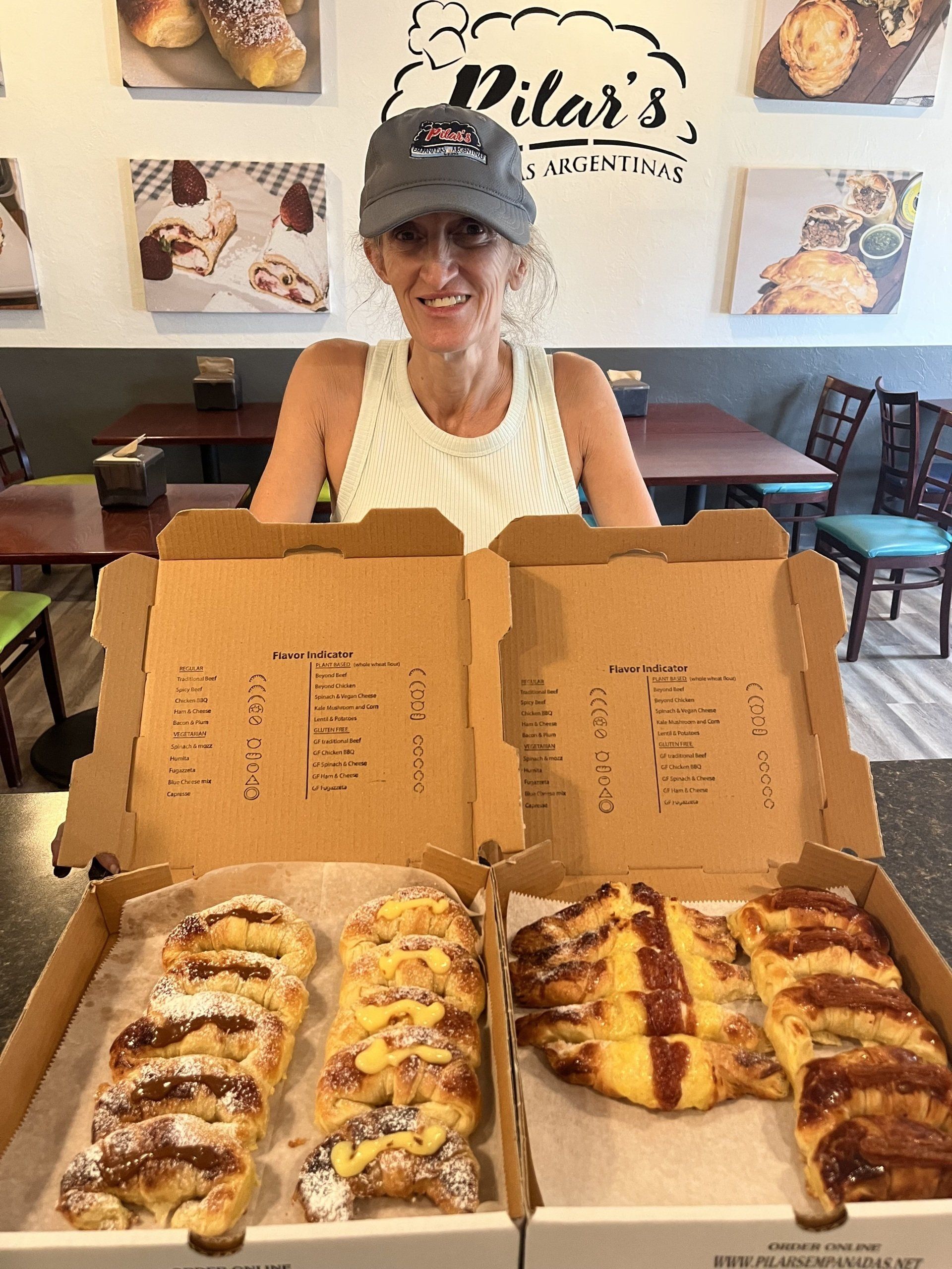 Woman with boxes of pastries at Pilar's Pastries. She wears a hat and smiles. Shop has tables and wall art.