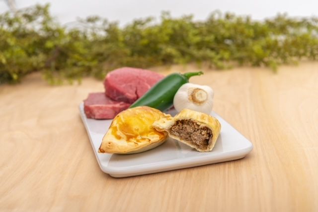 A cutting board with raw steak, a jalapeño, mushroom, and two empanadas on a wooden surface.