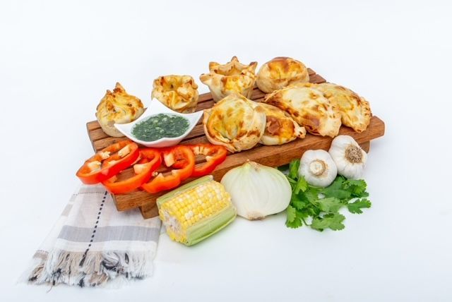 Assortment of baked pastries, vegetables, and dipping sauce on a wooden board; white background.