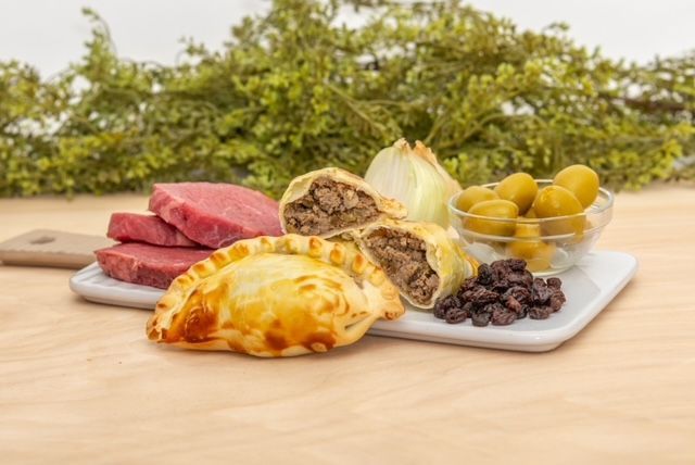 Meat empanadas, olives, meat slices, onion, and raisins on a white platter, wood background.