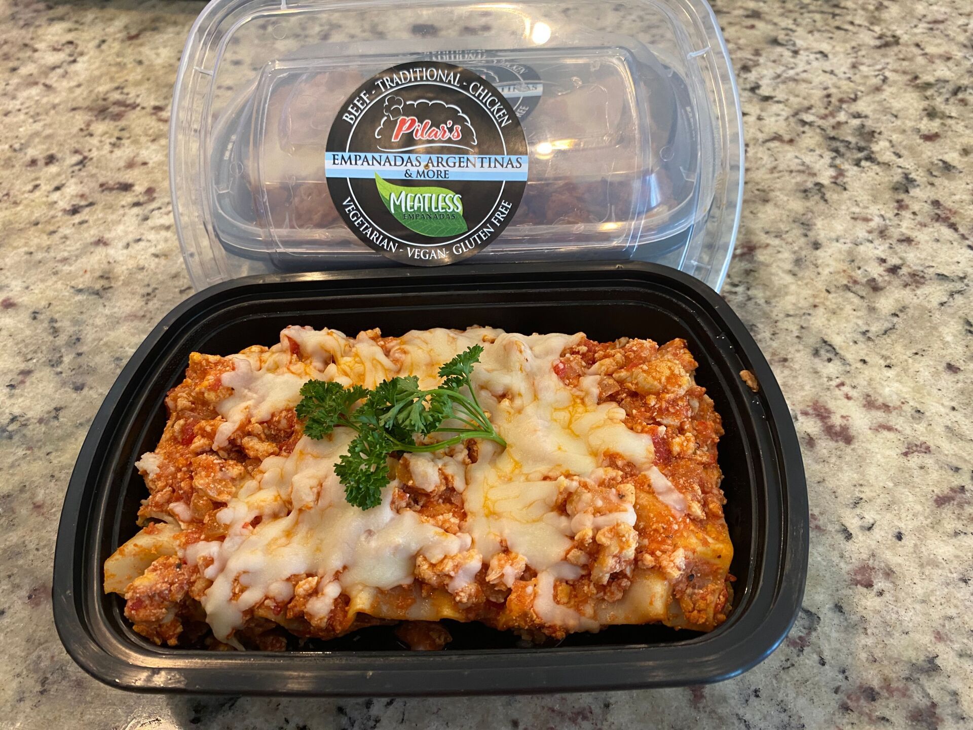 Lasagna in black container with a container of food behind it on a countertop.