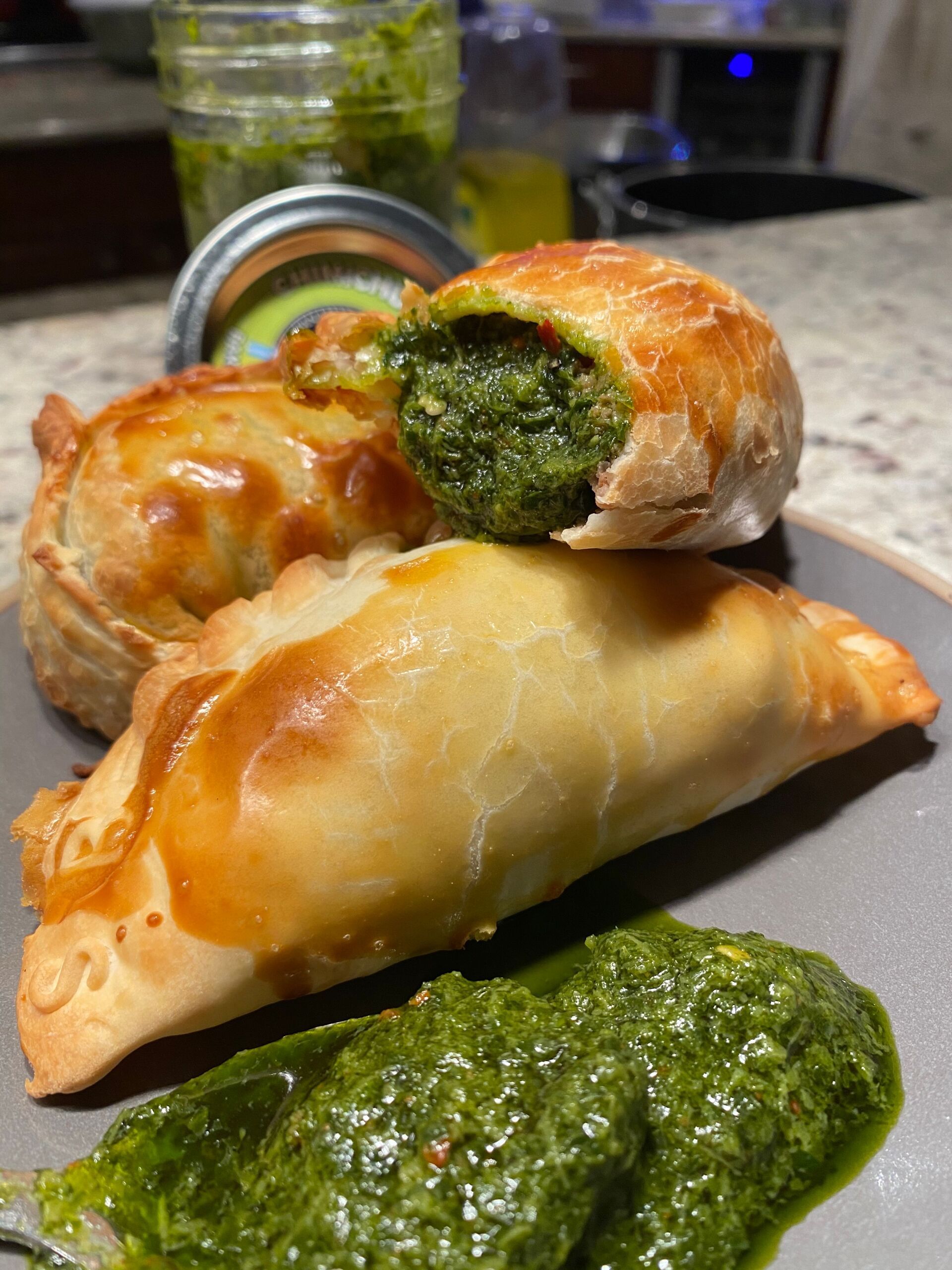 Golden-brown empanadas with green filling and pesto on a plate. A jar of pesto is visible in the background.
