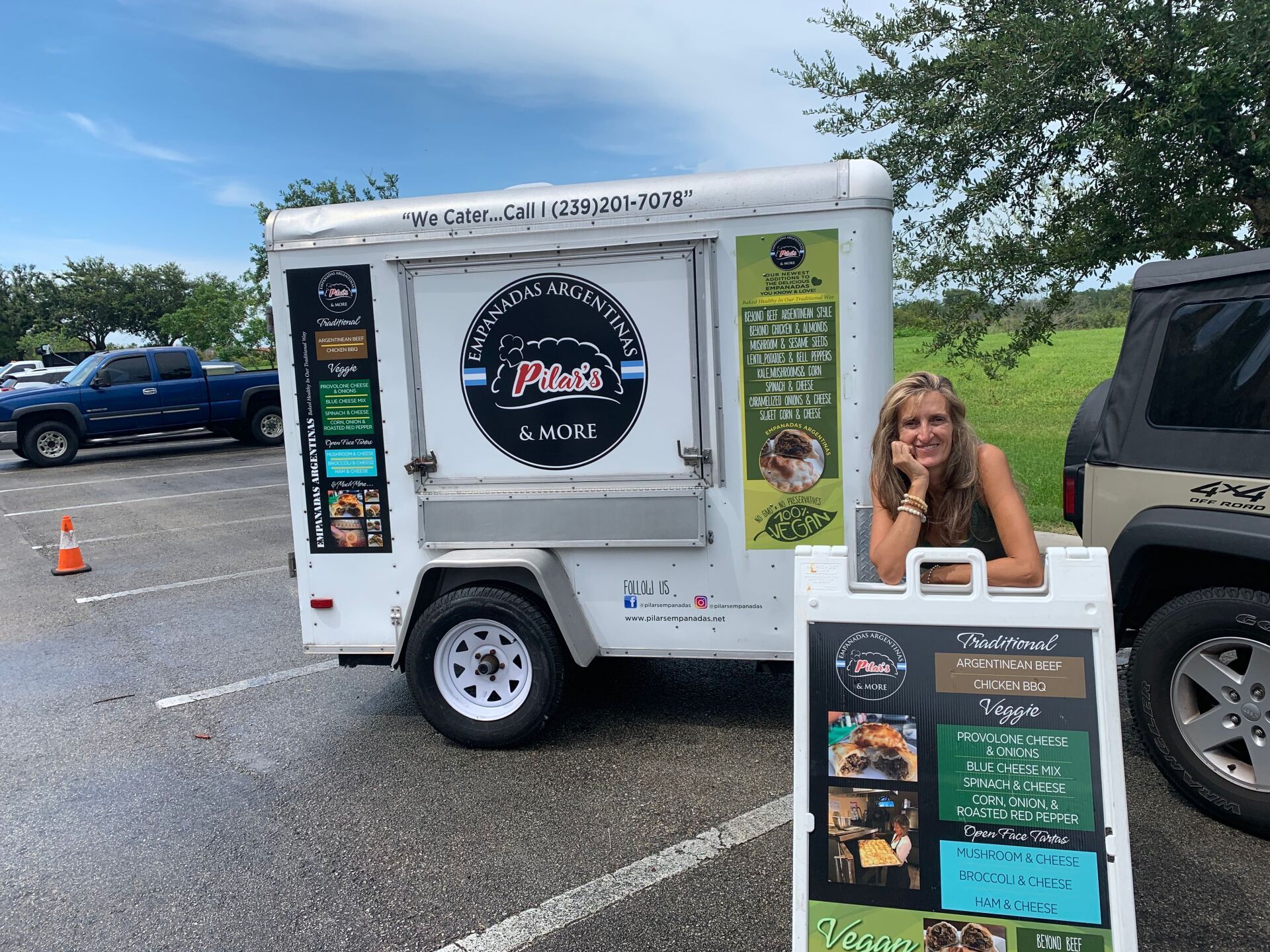 Woman stands by a food trailer, smiling. Sign in front, offers menu. Parking lot setting.