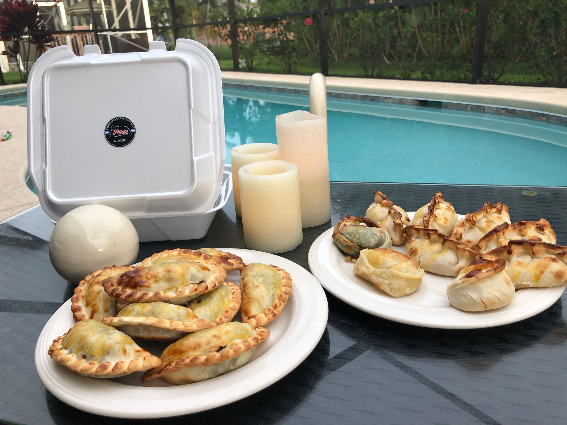 Empanadas, dumplings, candles, and a takeout container on a table beside a swimming pool.