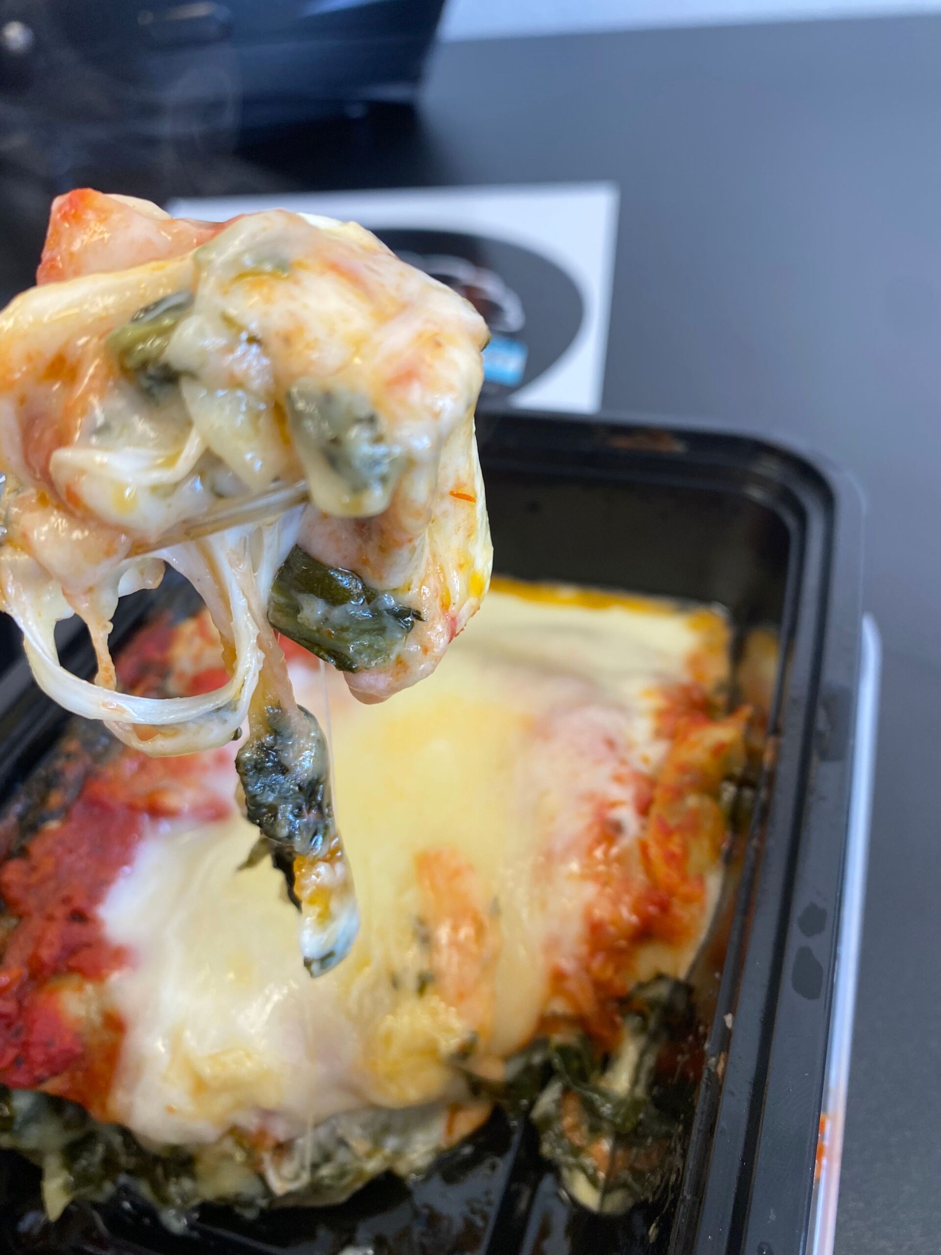 Lasagna with spinach and cheese, held up with melted cheese dripping from a black container on a table.