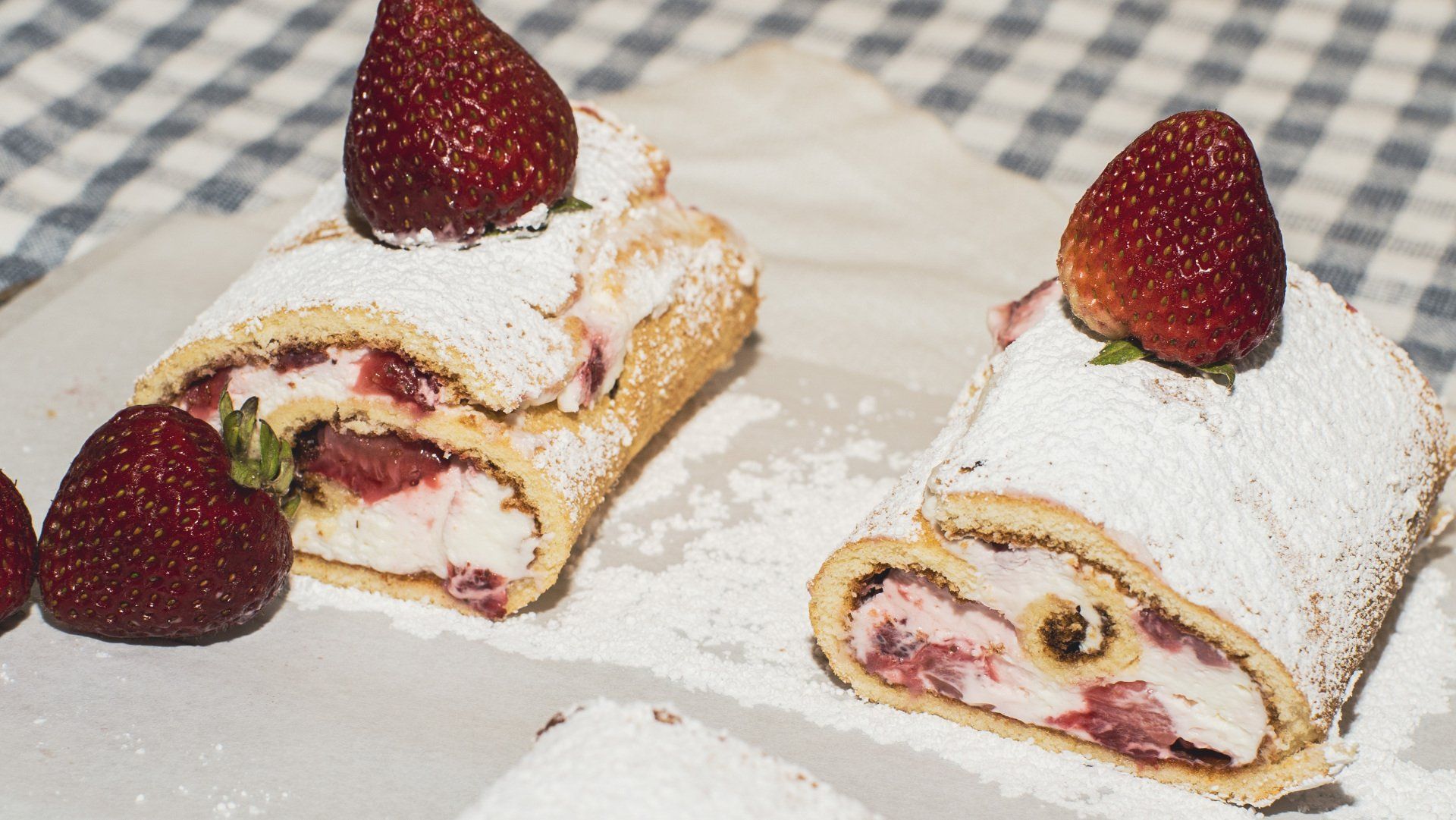 Two slices of strawberry cream roll topped with strawberries, dusted with powdered sugar, on parchment.