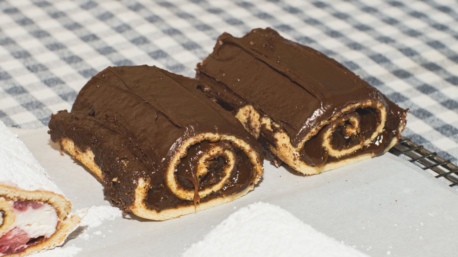 Two chocolate spiral pastries with chocolate frosting on a checkered tablecloth.