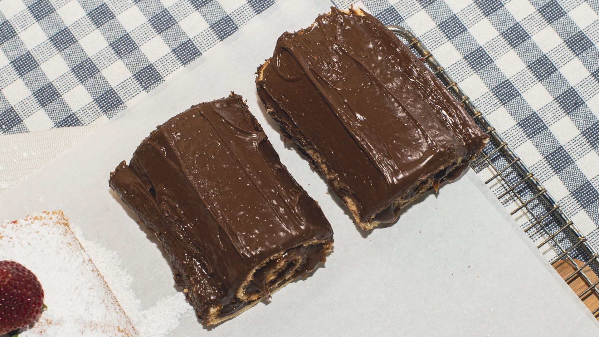 Two slices of chocolate roll cake with frosting on a white surface next to a checkered cloth.