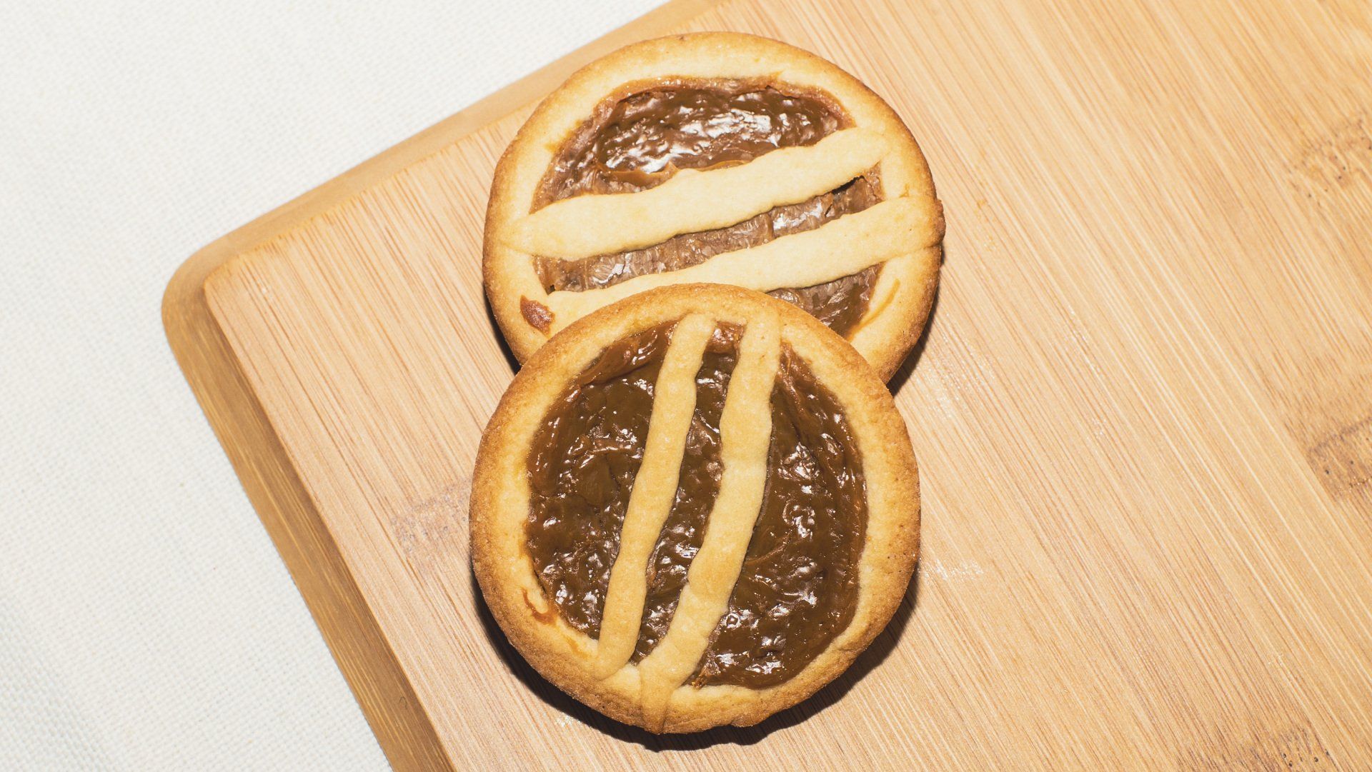 Two round, lattice-topped tarts filled with brown jam, on a wooden cutting board.