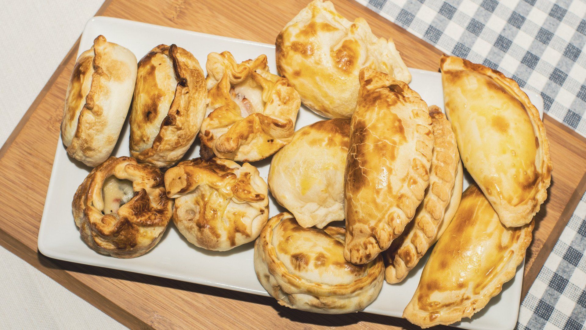Assorted golden-brown baked pastries on a white platter over a wooden board, with a checkered cloth in the background.