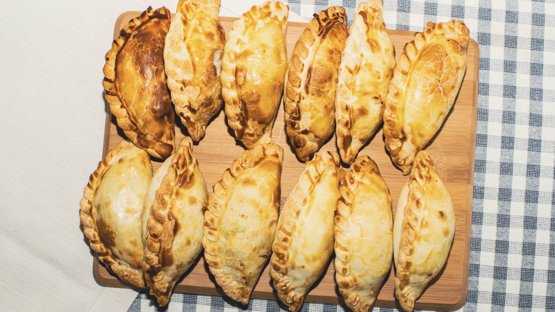 Golden-brown baked empanadas arranged on a wooden board, set on a checkered tablecloth.