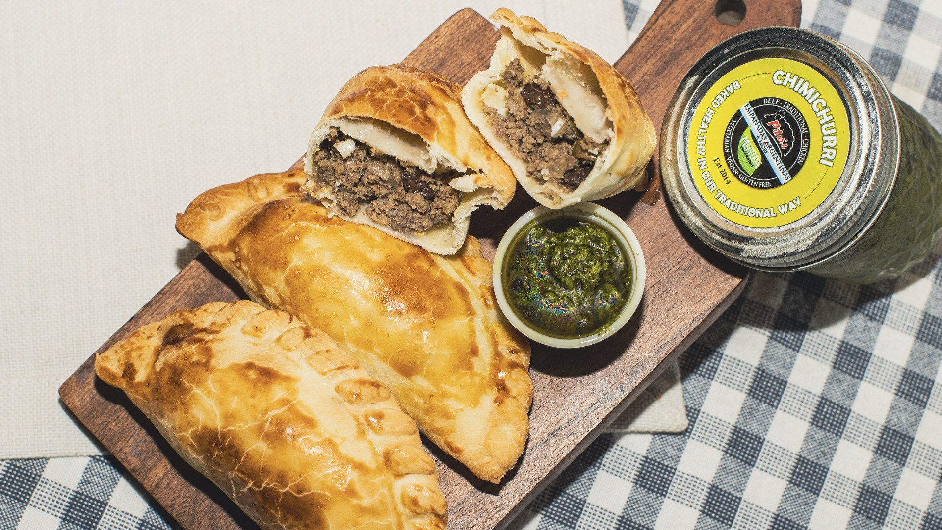 Empanadas on a wooden board with a small bowl of green sauce and a jar.