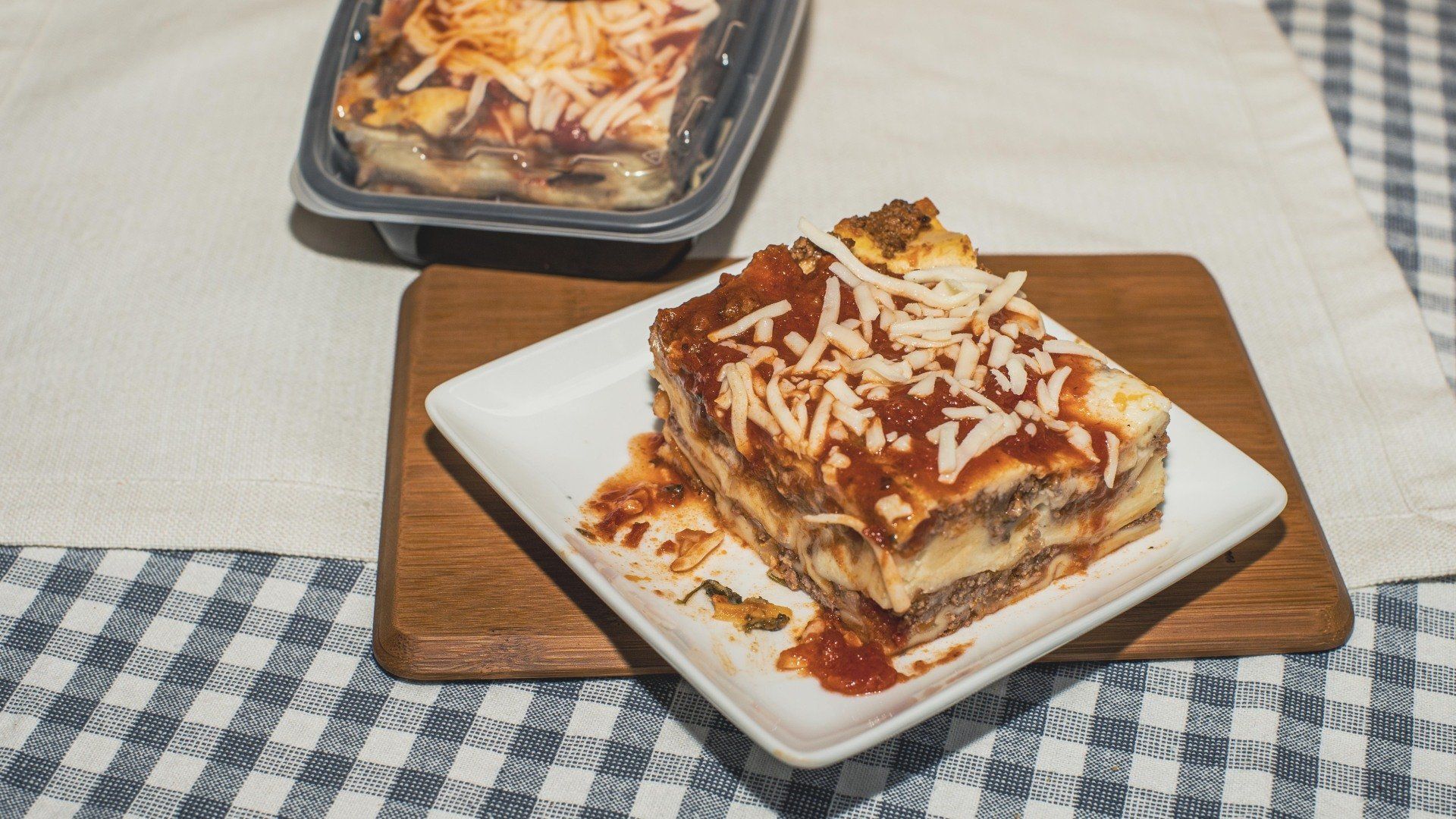 Lasagna on a white plate atop a wooden board, with another container of lasagna in the background on a checkered tablecloth.