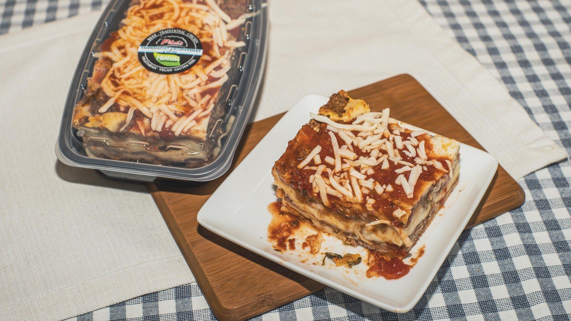 Lasagna in plastic container and a serving on a white plate atop a wooden board.
