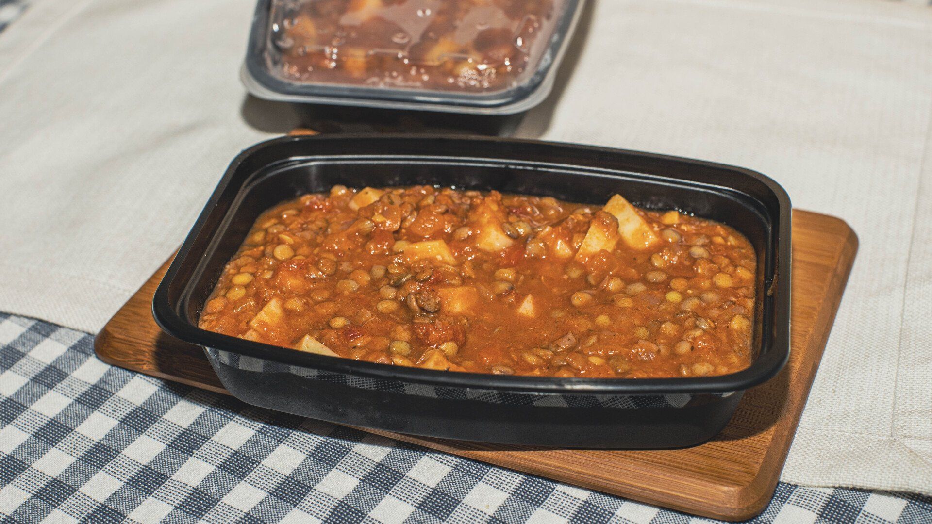 Lentil stew with potatoes in a black container on a wooden tray, another container in the background.