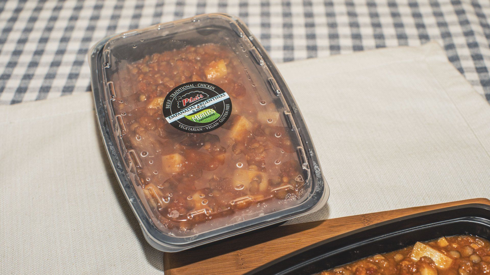 Plastic container of reddish food with a label, on a white cloth, checkered background.