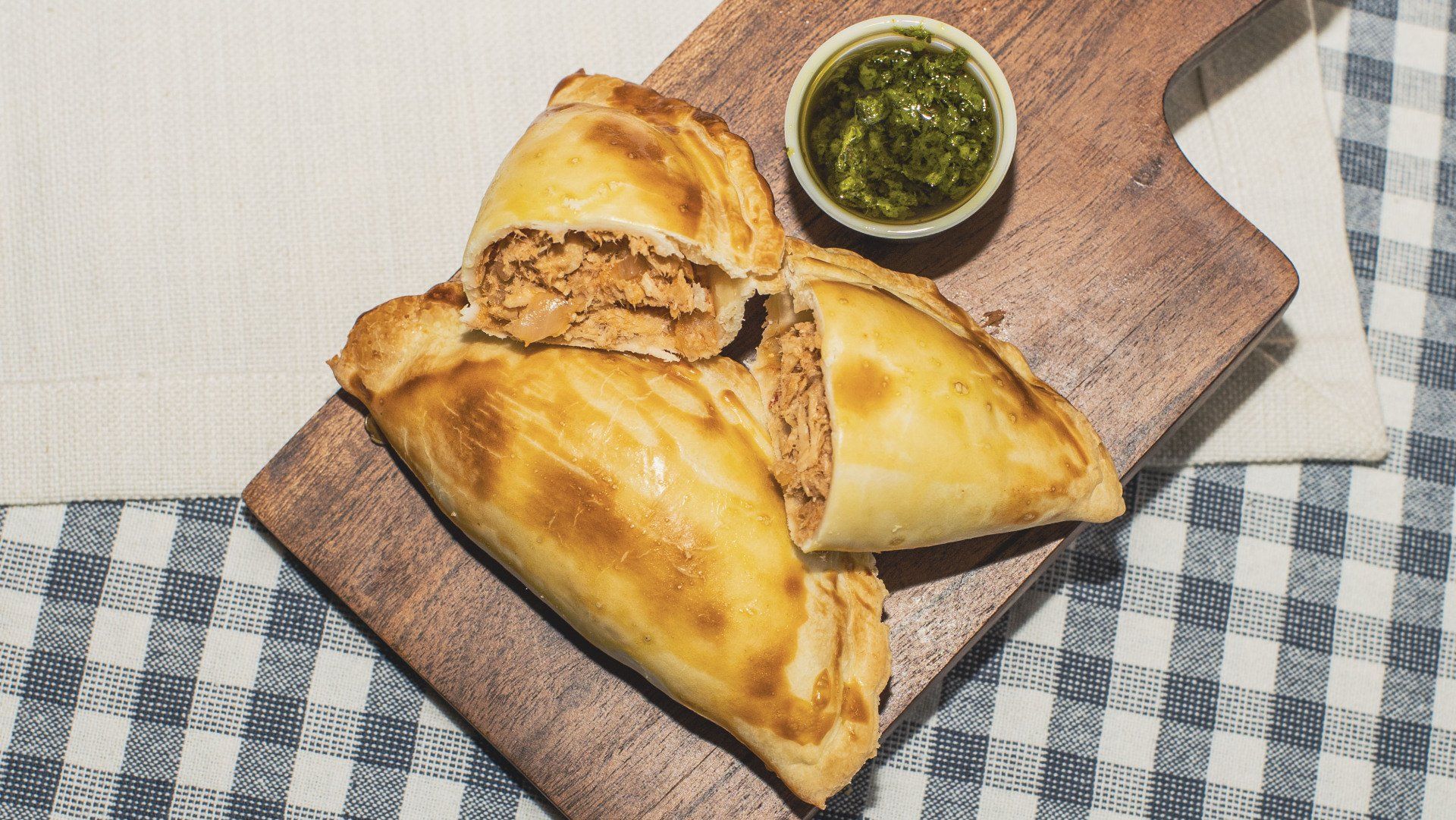 Chicken empanadas on a wooden board with a side of green sauce, on a checkered tablecloth.