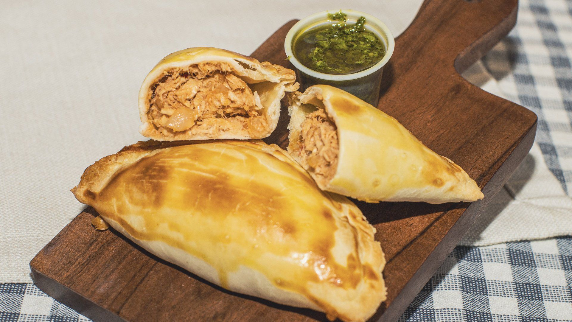 Empanadas on wooden board with green sauce, one cut open to show chicken filling.