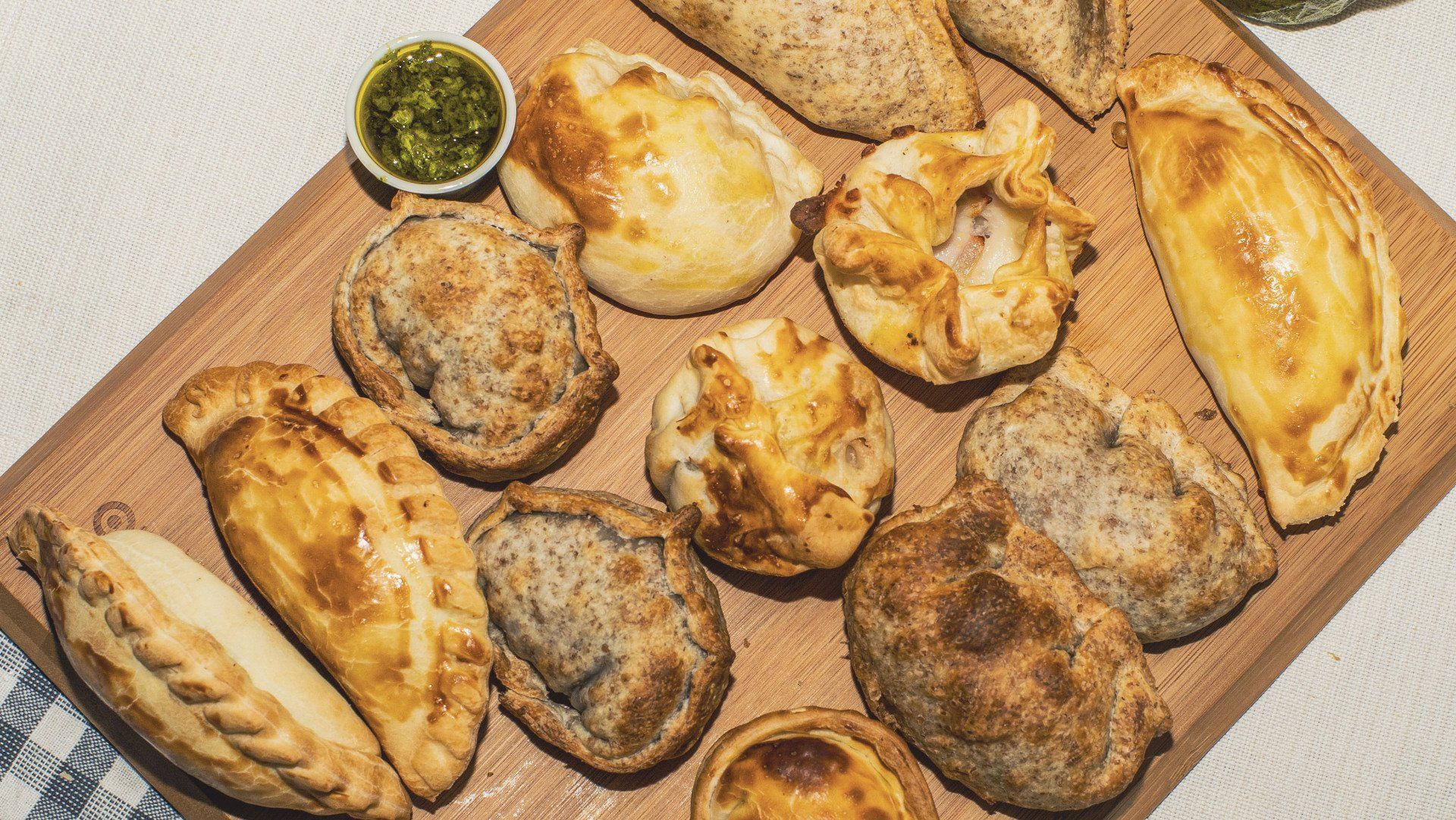Assortment of golden-brown empanadas on a wooden board, with a small bowl of green sauce.