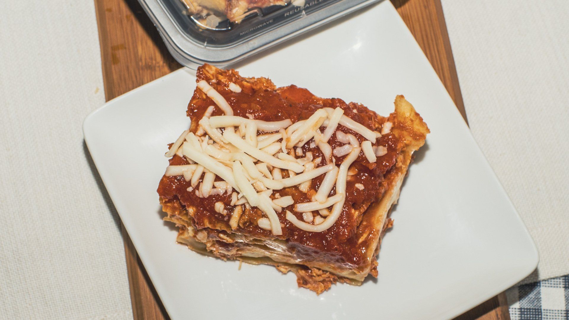 Lasagna slice on a white plate, topped with red sauce and shredded cheese, on a wood surface.