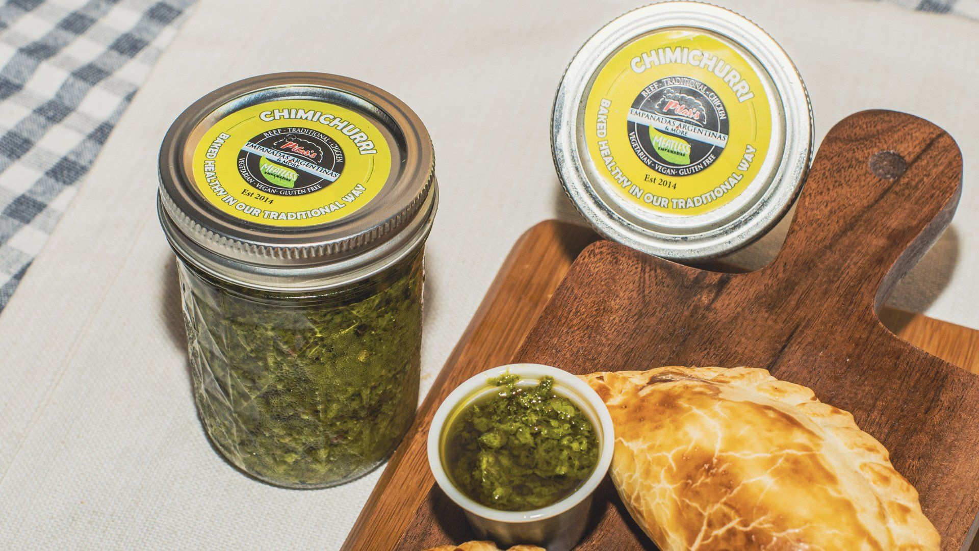 Two jars and a cup of green sauce, with empanada on wooden board.