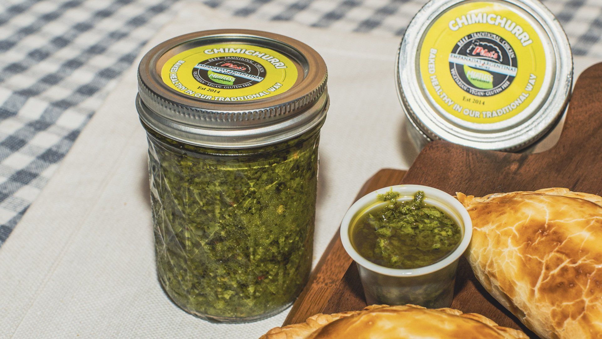 Jar of green chimichurri sauce with a small container, empanadas, and a lid on a wooden board.