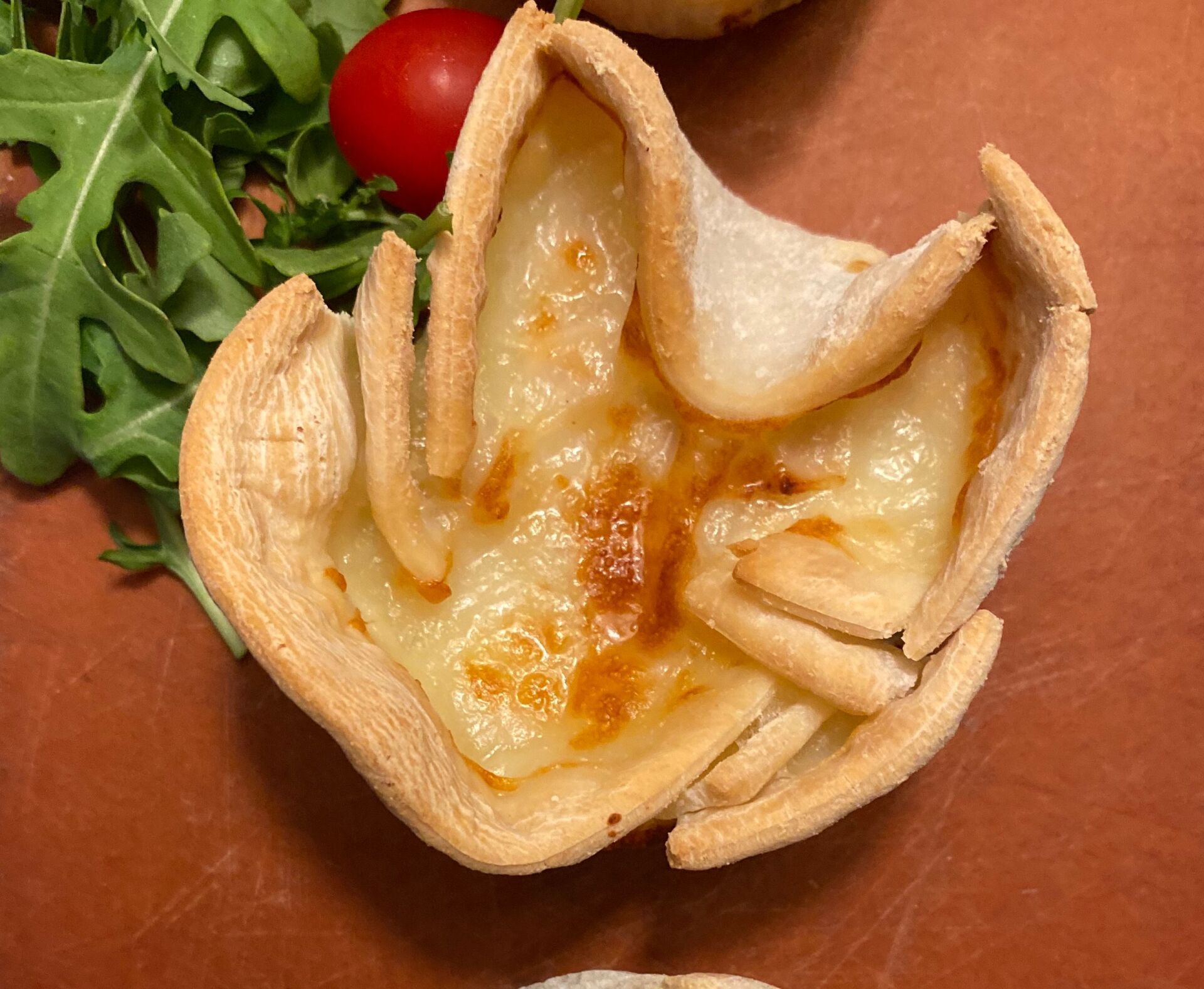 Savory tart with melted cheese, surrounded by folded crust, next to greens and a tomato.