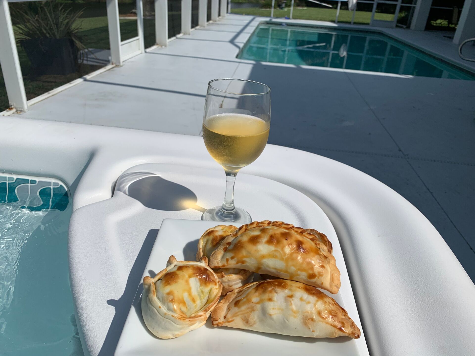 Empanadas and white wine on a serving tray next to a jacuzzi. Sunny, outdoor setting, pool in background.