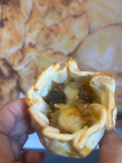 Hand holding an open-faced pastry with filling, a brown crust, and blurry background of similar pastries.