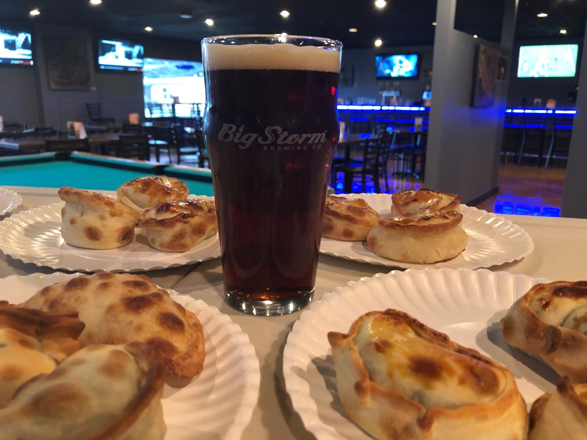 A pint of dark beer with empanadas on paper plates in a bar setting.