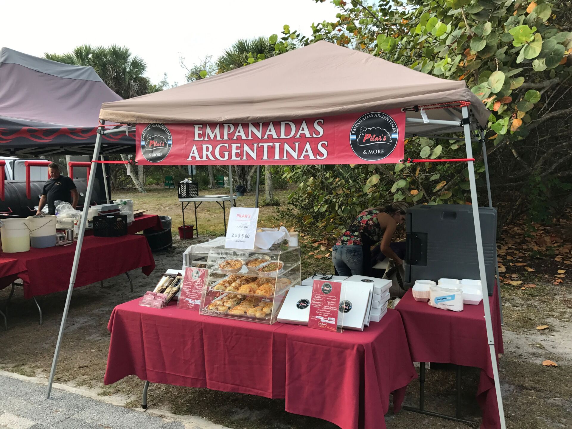 Argentine empanada stand at an outdoor event. Red tablecloths, beige canopy, sign.