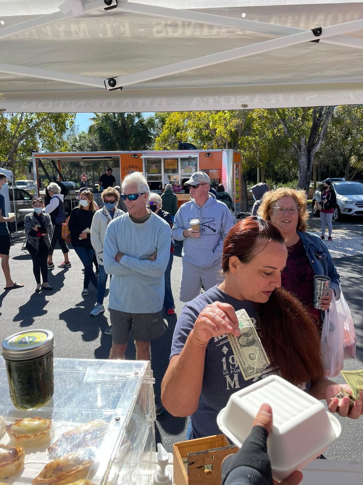 People line up at a market stall; a woman handles cash.