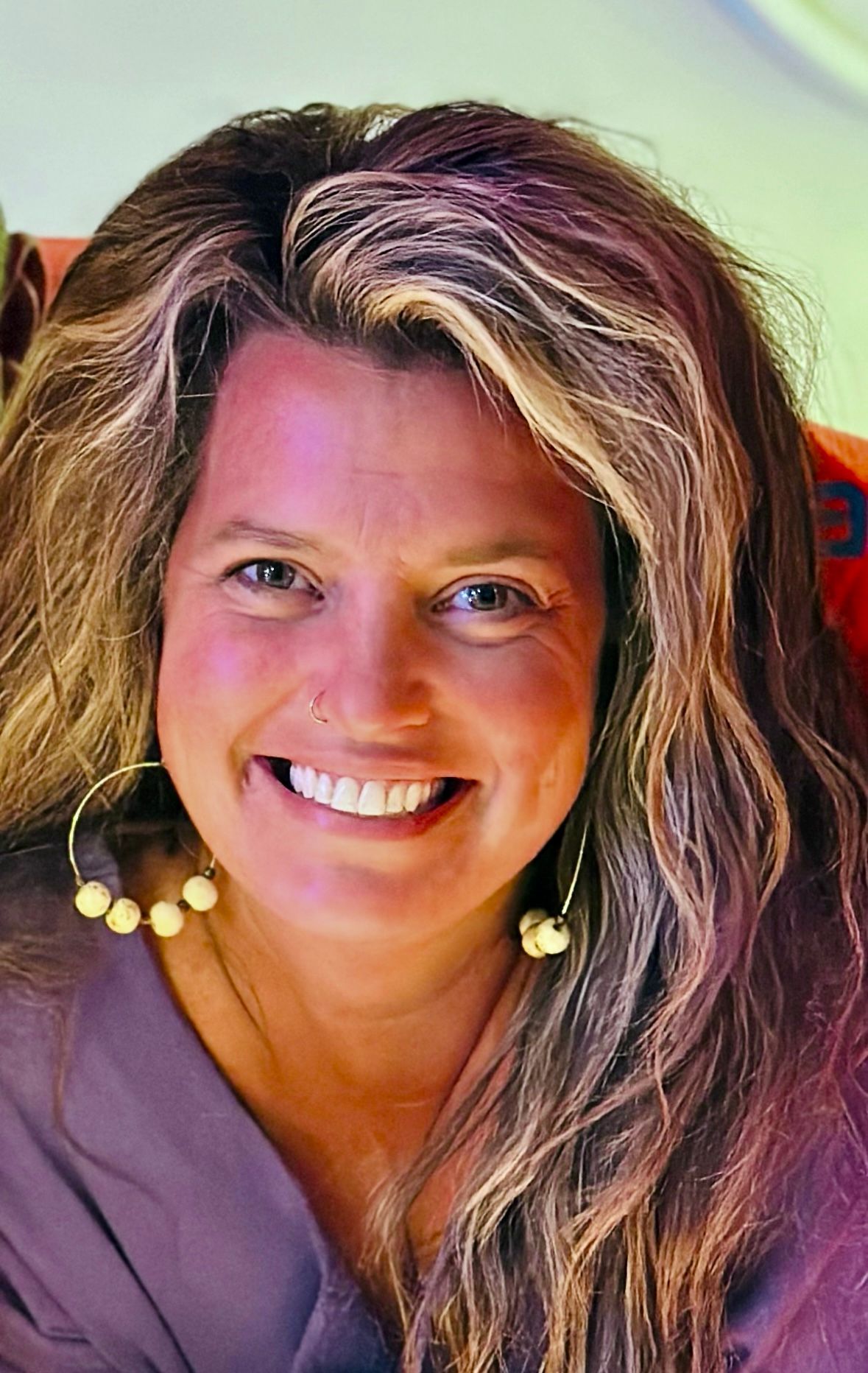Woman with long wavy hair smiles, wearing hoop earrings, nose ring, in a colorful setting.