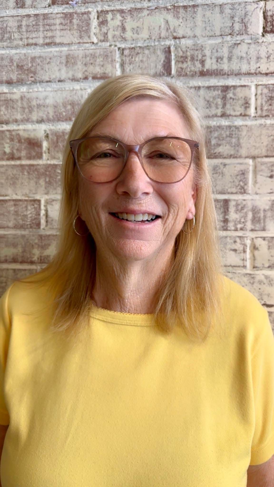 Woman wearing glasses and yellow shirt smiles in front of a brick wall.
