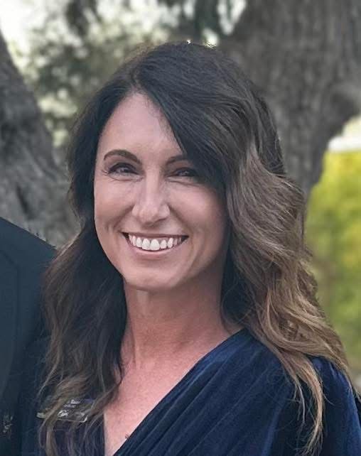 Woman with dark wavy hair smiles, wearing a navy blue top, outdoors.