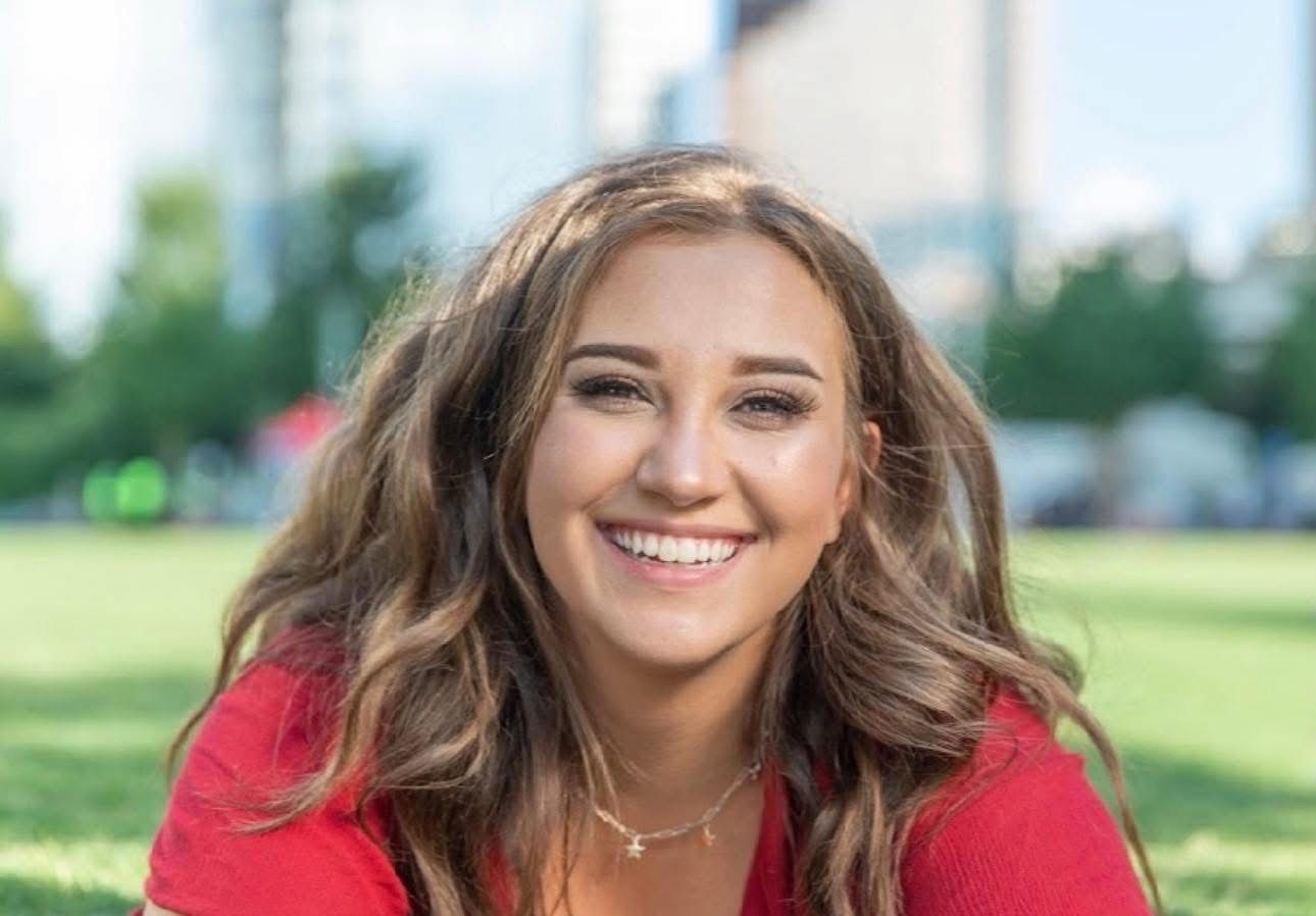 Woman with long wavy brown hair smiles at the camera, wearing a red top, lying on grass. Buildings in background.