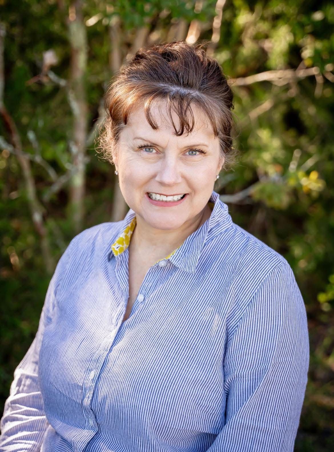 Woman smiling, wearing a blue and white striped shirt outdoors, with trees in the background.