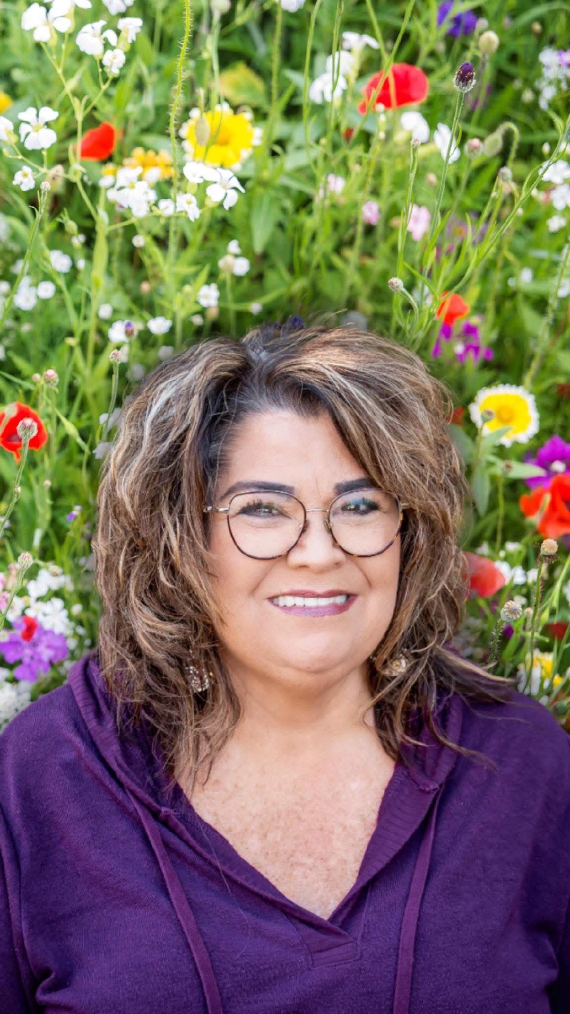 Woman with glasses smiles in front of colorful wildflowers; she wears a purple hooded sweatshirt.
