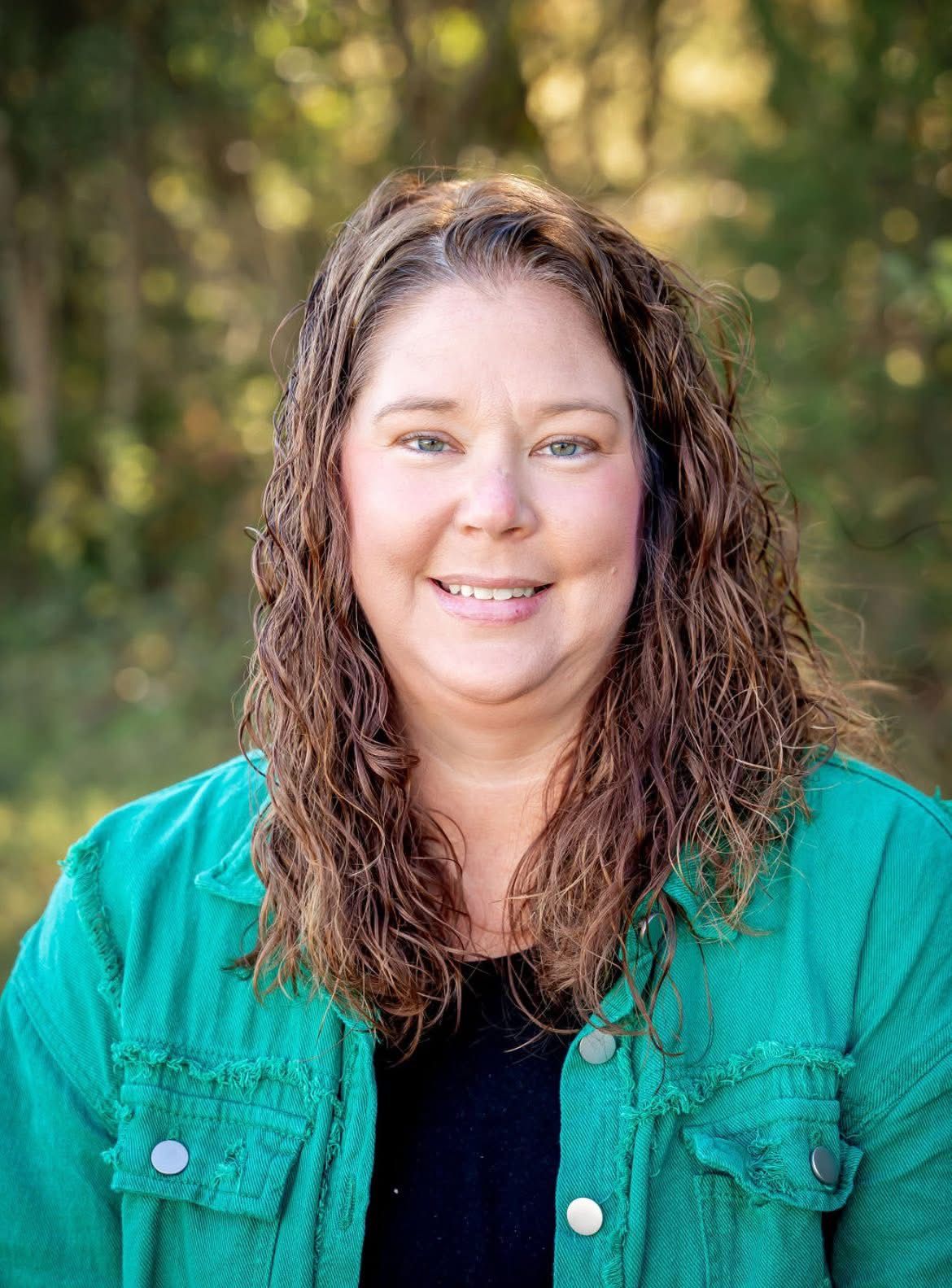 Woman with brown curly hair, smiling, wearing a green jacket over a black top, outdoors in front of blurred trees.