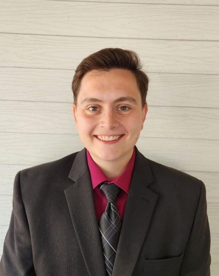 Man in suit, burgundy shirt, and patterned tie smiling in front of a light wood backdrop.