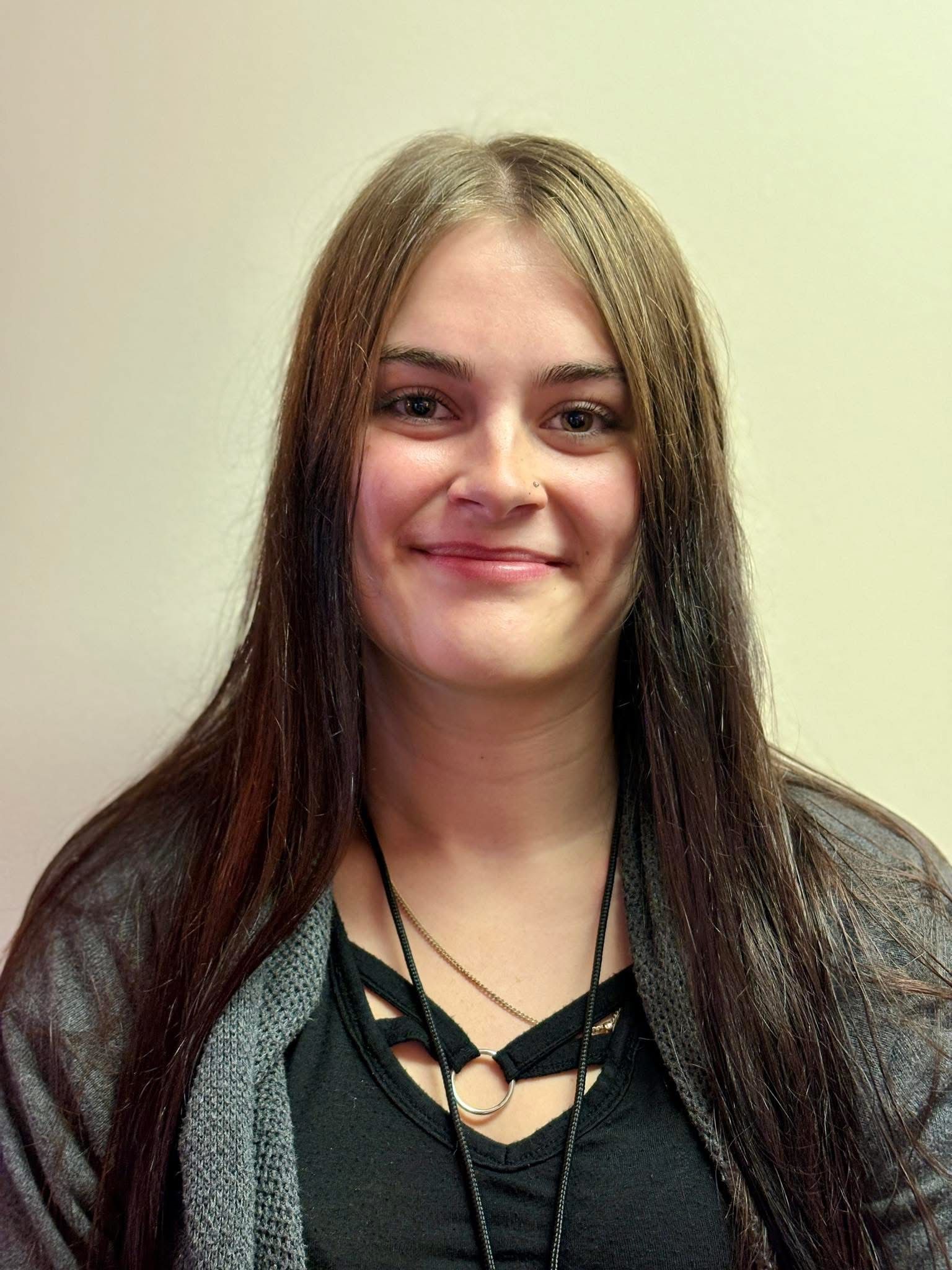 Woman with long brown hair smiling at the camera, wearing a black shirt and gray cardigan.