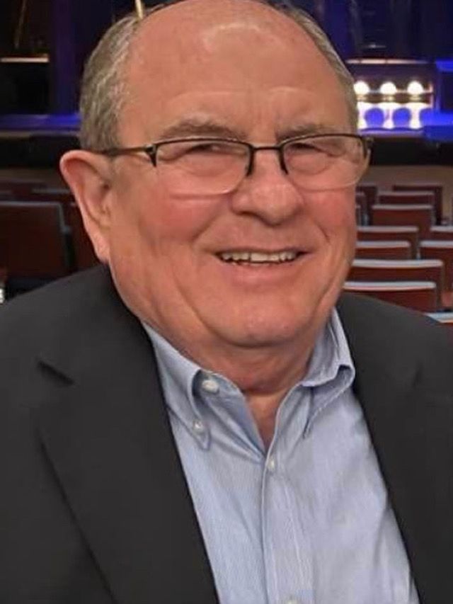 Man with glasses smiles, wearing a blue shirt and dark blazer. Background shows an auditorium.