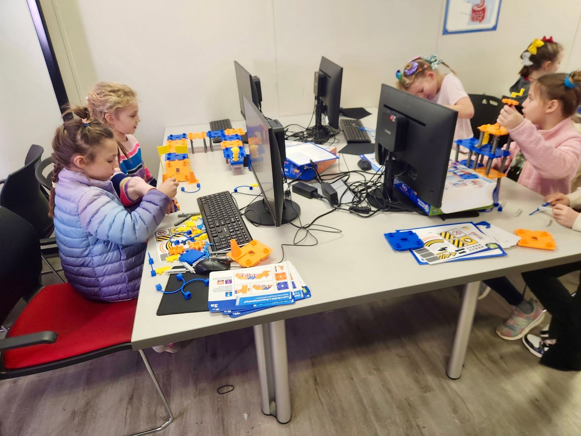Children building with construction toys at a table with computers in a classroom setting.