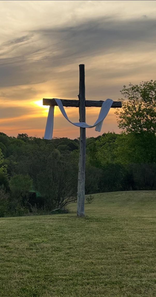 Wooden cross draped with white cloth, silhouetted against a colorful sunset.