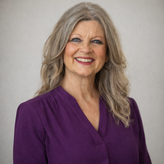 Woman with long, wavy gray hair smiles, wearing a purple top against a neutral background.
