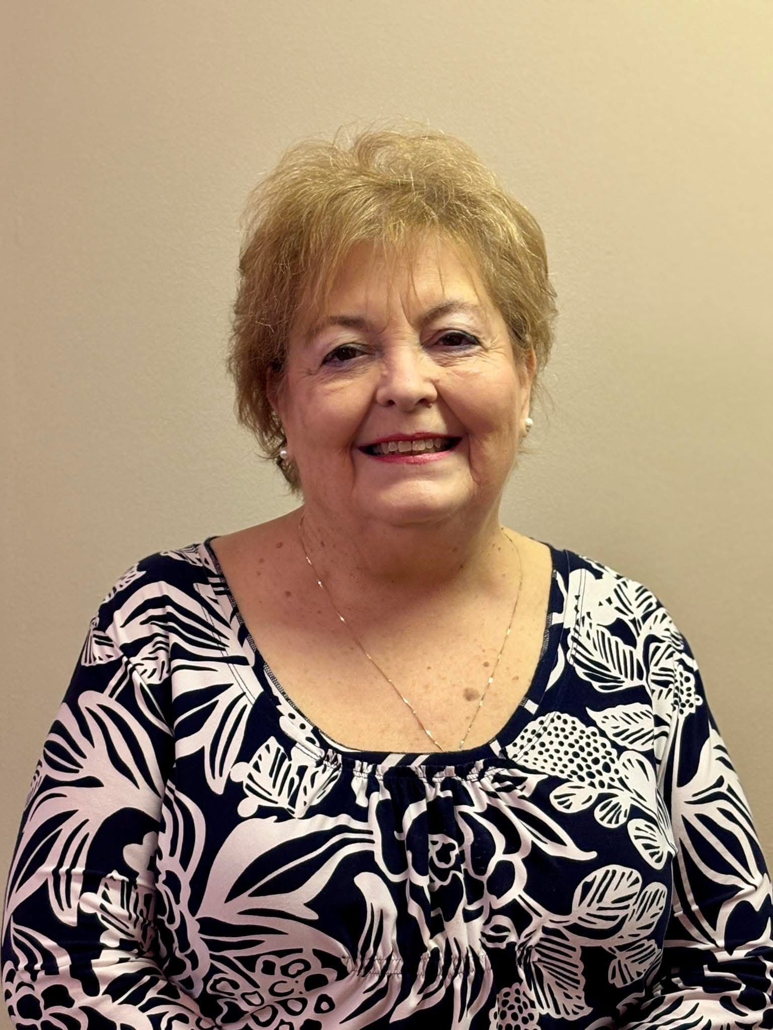 Woman with short, light-brown hair, smiling, wearing a navy and white patterned top, against a beige wall.