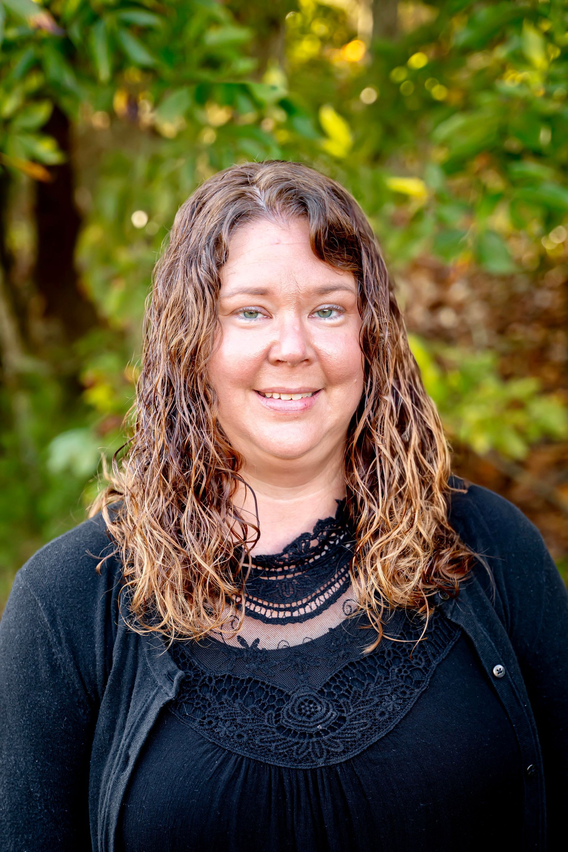 Woman with curly brown hair smiling, wearing a black top, against a blurred green background.